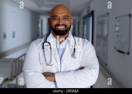 Porträt eines glücklichen birassischen männlichen Arztes, der Laborkittel und Stethoskop im Korridor des Krankenhauses trägt Stockfoto