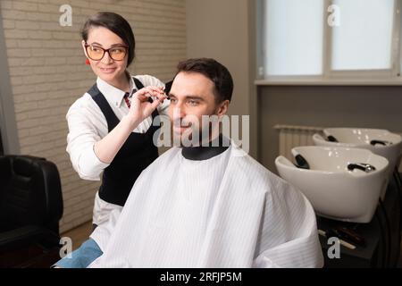 Ein junger Friseur schneidet einem Kunden die Haare mit einer Schere, neben einem speziellen Waschbecken zum Haarwaschen Stockfoto