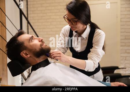 Weibliche Friseurin in Brille arbeitet mit einem Kundenbart, der Mann sitzt auf einem bequemen Stuhl Stockfoto