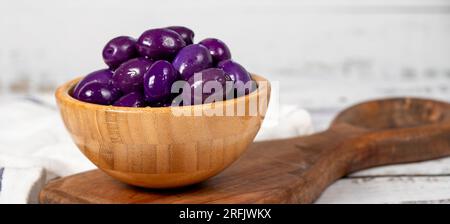Fermentierte Oliven mit Heidelbeeren in einer Holzschüssel. Lila Oliven auf weißem Holzhintergrund. Mediterrane Küche. Veganes Essen. Speicherplatz kopieren. Leerer Bereich für Text Stockfoto