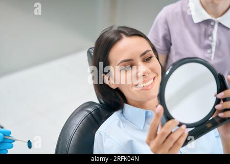 Frau im kieferorthopädischen Stuhl, die Spiegel betrachtet, die Qualität der Zahnaufhellung überprüft Stockfoto