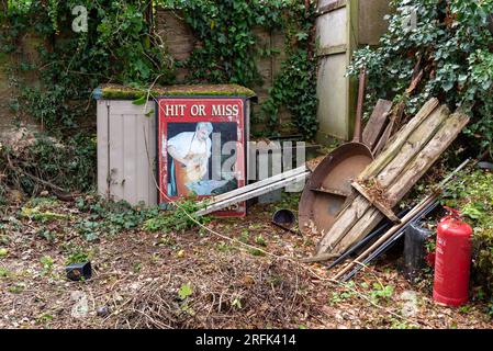 Auswahl an Artikeln, die draußen in einem Garten stehen und jetzt überwuchert sind. Alte Küchenpfanne, Feuerlöscher und ein Schild, das vielleicht von einem Unternehmen stammt. Stockfoto