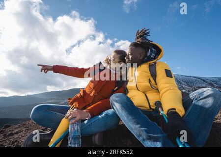 Ein junges multirassisches Paar, ein afrikanischer Mann und eine weiße Frau, die auf einem Berggipfel sitzt, der auf einen entfernten Horizont zeigt, verkleidet mit Outdoor-Ausrüstung. Stockfoto