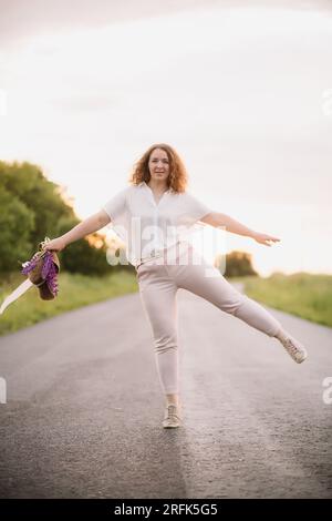 Eine junge Frau steht in einem weißen Hemd auf der Straße mit lila und pinkfarbenen Lupinen. Schöne junge Frau mit lockigem Haar und Lupinen-Strauß. Sonnenuntergang oder Sonnenaufgang Stockfoto