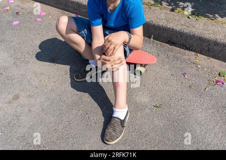 Abrieb am Bein eines Kindes, wenn es beim Skateboard-Fahren auf einen Asphaltpfad fällt. Der Junge ist beim Skateboard-Fahren gestürzt. Extremsport. Stockfoto