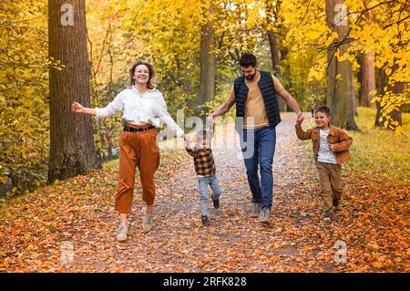 Glückliche Familie: Mutter, Vater und zwei Söhne laufen durch den Herbstpark und halten Händchen. Der Boden ist mit gelben Blättern bedeckt. Familientagskonzept Stockfoto