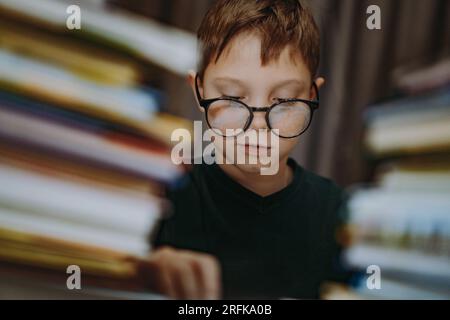 Süßer, weißer Junge, der eine Brille trägt und den Kopf mit einem Buch bedeckt. Ein fröhlicher Junge, der hinter Bücherhaufen guckt. Bild mit selektivem Fokus und Rauscheffekt Stockfoto