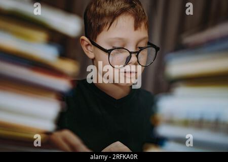 Süßer, weißer Junge, der eine Brille trägt und den Kopf mit einem Buch bedeckt. Ein fröhlicher Junge, der hinter Bücherhaufen guckt. Bild mit selektivem Fokus und Rauscheffekt Stockfoto