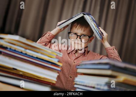 Süßer, weißer Junge mit Brille, der den Kopf mit einem Buch bedeckt. Ein fröhlicher Junge, der hinter Bücherhaufen guckt. Bild mit selektivem FO Stockfoto