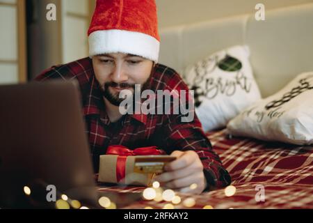 ein weißer, gutaussehender Mann in einem roten karierten Flanellhemd im weihnachtsmann, der mit einem Laptop auf dem Bett herumhutscht, sich online unterhält oder Pres kauft Stockfoto