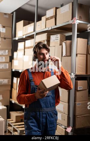 Lagerarbeiter in einem schnurlosen Headset, der in einem Lager arbeitet, trägt eine Box, während er mit Waren an den Regalen vorbeigeht. Stockfoto