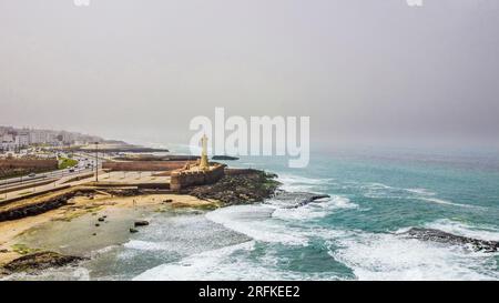 Drohnenansicht von Rabat, Marokko. Felsige Atlantikküste mit Leuchtturm, Wohngebäuden und Autobahn im Hintergrund, Nebel im A Stockfoto