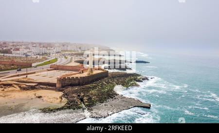 Drohnenansicht von Rabat, Marokko. Felsige Atlantikküste mit Leuchtturm, Wohngebäuden und Autobahn im Hintergrund, Nebel im A Stockfoto