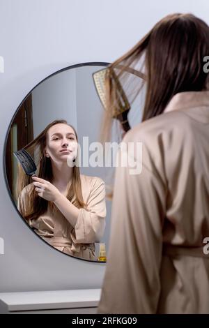 Hübsche Frau, Die Ihr Schönes Haar Mit Einer Bürste Kämmt, Während Sie Im Badezimmer Neben Dem Spiegel Steht, Attraktive Junge Dame, Die Auf Ihr Spiegelbild Schaut Und Lächelt Stockfoto