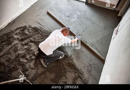 Mann glättet und nivelliert die Oberfläche mit geradem Schneidmesser, während der Boden in der Wohnung gehoben wird. Draufsicht eines Bauarbeiters, der die Estriche auf dem mit Sandzement-Mischung bedeckten Boden platziert. Stockfoto