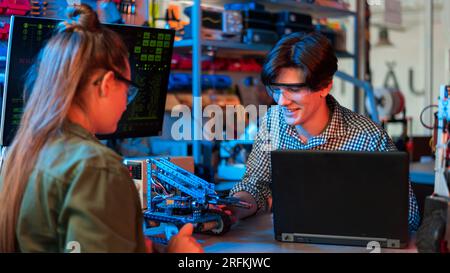 Teenager, die Roboterexperimente in einem Labor machen. Junge und Mädchen in Schutzbrille arbeiten mit einem Roboter, Laptop auf dem Tisch. Rot und blau illumi Stockfoto