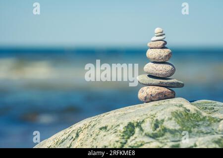 Balancieren von Steinen auf einem großen Felsen mit Seegras am Strand, mit blauen Wasserwellen im Hintergrund, Ostsee, Olando Kepure, Litauen Stockfoto
