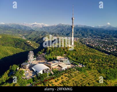 Drohnenaufnahme des Koktobe Hill Park mit Riesenrad, einem Wahrzeichen der Stadt Almaty, mit dem der hohe Fernsehturm vor den Schneebirgen Kasachstans steht Stockfoto