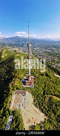 Vertikale Luftdrohnenaufnahme des Hochfernsehturms der Stadt Symbol Almaty und Parken auf dem Koktobe-Hügel vor den Schneebirgen Kasachstans Stockfoto