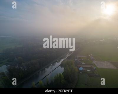 Tauchen Sie ein in die bezaubernde Schönheit eines mystischen Vormittags, während die Kamera über einen fesselnden Blick aus der Vogelperspektive auf einen Fluss, ein Bauernfeld und eine Waldlandschaft schwenkt. Unter der sanften Umarmung des Morgennebels schlängelt sich der ruhige Fluss durch die üppigen grünen Felder und den majestätischen Wald. Wenn die Sonne aufgeht, durchdringen die goldenen Strahlen den Nebel und werfen ein warmes und ätherisches Licht auf die Landschaft. Dieser Schwenkvorgang fängt die Essenz der Ruhe und den Reiz des Erwachen der Natur ein. Erleben Sie die ruhige Schönheit und das fesselnde Ambiente dieser magischen Morgenszene, wo das Zusammenspiel von Nebel stattfindet Stockfoto