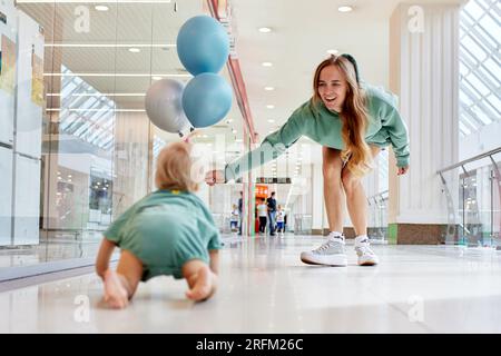 Fröhliche lächelnde Mutter mit ihrem Baby, das auf dem Boden krabbelt und farbige Ballons. Junge, hübsche mutter und ihre kleine Tochter in grünen Klamotten laufen um mich herum Stockfoto