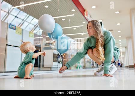 Fröhliche lächelnde Mutter mit ihrem Baby, das auf dem Boden krabbelt und farbige Ballons. Junge, hübsche mutter und ihre kleine Tochter in grünen Klamotten laufen um mich herum Stockfoto