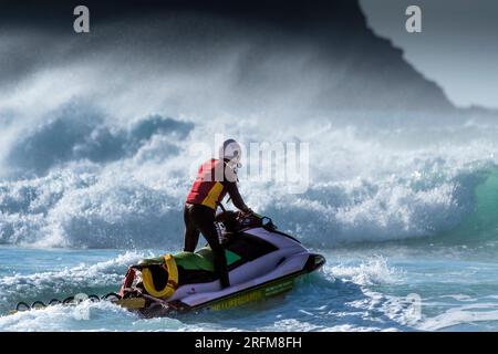 Ein RNLI-Rettungsschwimmer, der im Dienst auf einem Jetski in Fistral in Newquay in Cornwall in England im Vereinigten Königreich sitzt. Stockfoto