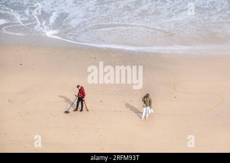 Ein Metalldetektorist sucht und eine Frau, die ein Handy benutzt, in Tolcarne Beach in Newquay in Cornwall, Großbritannien. Stockfoto