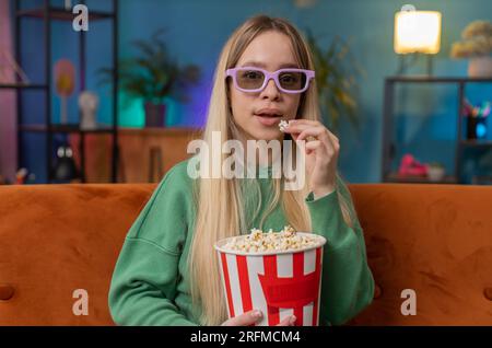 Aufgeregte Frau mit 3D-Brille sitzt auf dem Sofa und isst Popcorn-Snacks und sieht sich interessante TV-Serien-Sportspiele, Filme und Online-Social-Media-Filme zu Hause an. Ein Mädchen, das Unterhaltung genießt Stockfoto