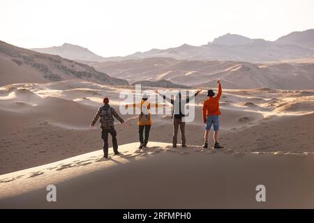 Vier glückliche Touristen mit Rucksäcken stehen auf Sanddünen mit offenen Armen und blicken auf den Sonnenuntergang Stockfoto