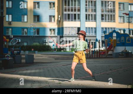 Ein süßes Mädchen im Schulalter bläst im Hof eines mehrstöckigen Gebäudes im Sommer große Seifenblasen. Sommerkonzept, Spiele im Freien Stockfoto