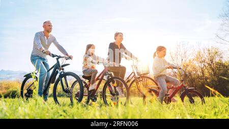 Lächelnde Eltern und Mutter mit zwei Töchtern während der Sommerradtour im Freien. Sie genießen die Zusammengehörigkeit auf grüner Wiese mit hohem Gras. Glückliches Parenthoo Stockfoto