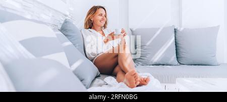 Schöne Barfuß Frau gekleidet weißes Hemd entspannen auf Luxus-Terrasse mit Espresso-Tasse Terrasse auf dem bequemen Sofa Kissen und genießen Sie Mittag ti Stockfoto