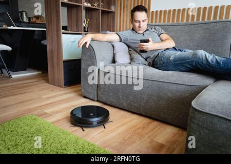 Roboter-Staubsauger Reinigung des Wohnzimmers. Mann genießen Ruhe, sitzen auf dem Sofa zu Hause. Fernbedienung des Roboter-Staubsaugers über das Smartphone. Smart t Stockfoto