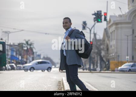 Latino-Mann im Anzug und Rucksack, der die Straße überquert, in einer Stadt mit Kopierraum. Stockfoto