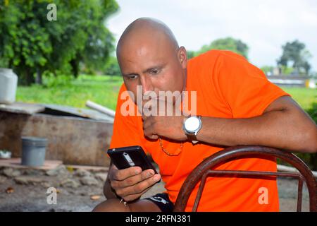 Süßes Glatzkopf-Model mit orangefarbenem T-Shirt, fröhliche Männer, die auf Handy schauen, Bauernhof-Hintergrund Stockfoto