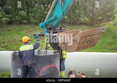 Arbeiter arbeiten auf der Baustelle. Ein Hydraulikbagger verwendet eine Kette, um ein Betonrohr anzuheben und einen Regenwasser- oder Kanalisationssammler zu installieren Stockfoto