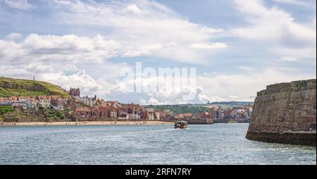 Whitby Hafeneingang. Die Gebäude liegen am Meer und die Leute sind an einem Sandstrand. Ein Touristenboot ist im Wasser und eine Steinmauer im Vordergrund Stockfoto