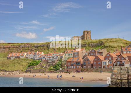 Die Leute sind an einem Sandstrand an einem Hügel mit roten Dachhäusern. Eine Kirche befindet sich auf dem Hügel und ein Himmel mit Lichtwolke ist darüber. Stockfoto