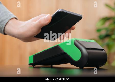 Nahaufnahme der Hand eines Mannes, der sein Smartphone in der Nähe des nfc-Zahlungsterminals hält. Kunde nutzt Telefon für kontaktlose Bezahlung im Geschäft während des Einkaufs, bargeldlose Technologie und NFC-Zahlungskonzept Stockfoto
