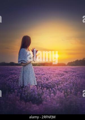 Das Seitenporträt einer jungen Frau in Kleidern steht auf dem violetten Lavendelfeld mit Blick auf den wunderschönen Sonnenuntergang. Natürliche Sommerdämmerung Stockfoto