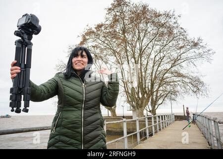 Eine junge venezolanische Vloggerin aus latina, die lächelt und ihre Kamera hält und ein Video mit Erklärungen und Gesprächen macht. Sie steht auf einem Pier des Hafens von olivos in Bu Stockfoto