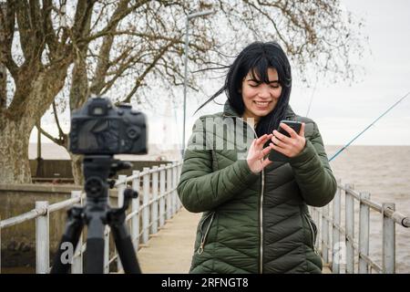 Eine junge lateinische Frau, die mit einem Lächeln Inhalte erstellt, Fotos und Videos im Freien aufnimmt und ihre Digitalkamera mit einer App auf ihrem Smartphone, Technol, steuert Stockfoto