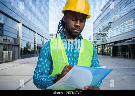 Vorderansicht eines erwachsenen männlichen Bauingenieurs aus afrikanischer ethnischer Herkunft, trägt einen gelben Schutzhelm, eine reflektierende Weste und Freizeitkleidung, steht mit Gebäuden Stockfoto