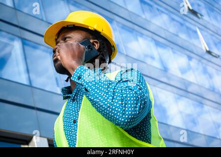 Porträt eines schwarzen männlichen Ingenieurs, der einen gelben Sicherheitshelm und eine reflektierende Weste trägt, draußen steht, telefoniert und sich umsieht, mit Comm Stockfoto