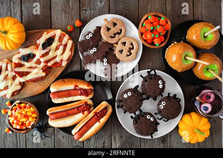 Halloween-Party mit Esstischszene vor rustikalem Holzhintergrund. Draufsicht. Gruselige Mumienpizzen, Finger-Hotdogs, Karameläpfel, Cupcakes, Süßigkeiten, Stockfoto