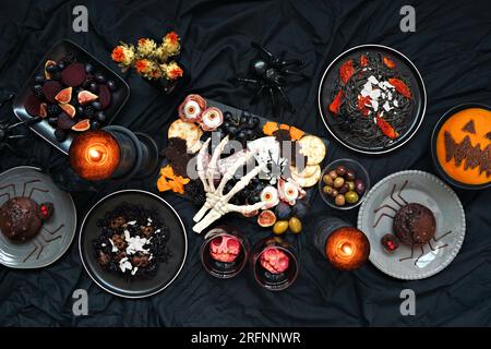 Halloween-Dinner-Party-Tischszene auf dunklem schwarzen Hintergrund. Ansicht von oben nach unten. Gruselige Wurstplatte, schwarzes Risotto und Pasta, Spinnenkuchen, Jack o Stockfoto