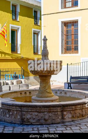 Villajoyosa, Spanien, mittelalterlicher Steinbrunnen in der Mitte einer kleinen Parkette. Der Boden des Platzes ist aus Kopfsteinpflaster. Stockfoto