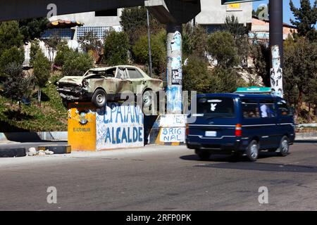 Minibus fährt an einem Unfallauto vorbei, das auf einem Sockel mitten auf der Autobahn steht. Fahrer werden gewarnt, nicht zu trinken, fahren und vorsichtig fahren zu dürfen, La Ceja, El Alto, Bolivien Stockfoto