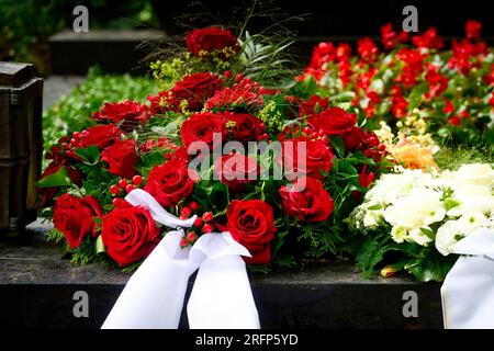 Ein Strauß von Grabblumen aus roten Rosen mit weißem Trauerbogen auf einem Grab nach einer Beerdigung Stockfoto